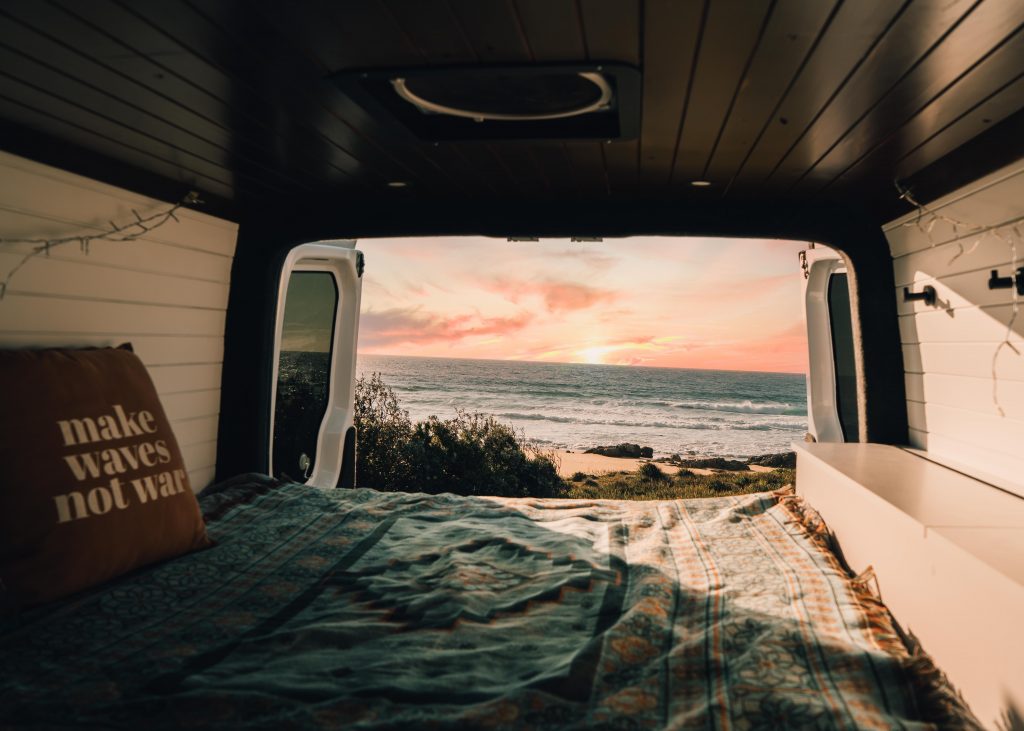 We are looking out the back of a van. It is a pink sunset over a crystal blue sea. A pillow saying Make Waves Not War can be seen atop a blue blanket. This person has chosen a simple van life with no kitchen or bed visible. Just the beautiful, natural scenery.