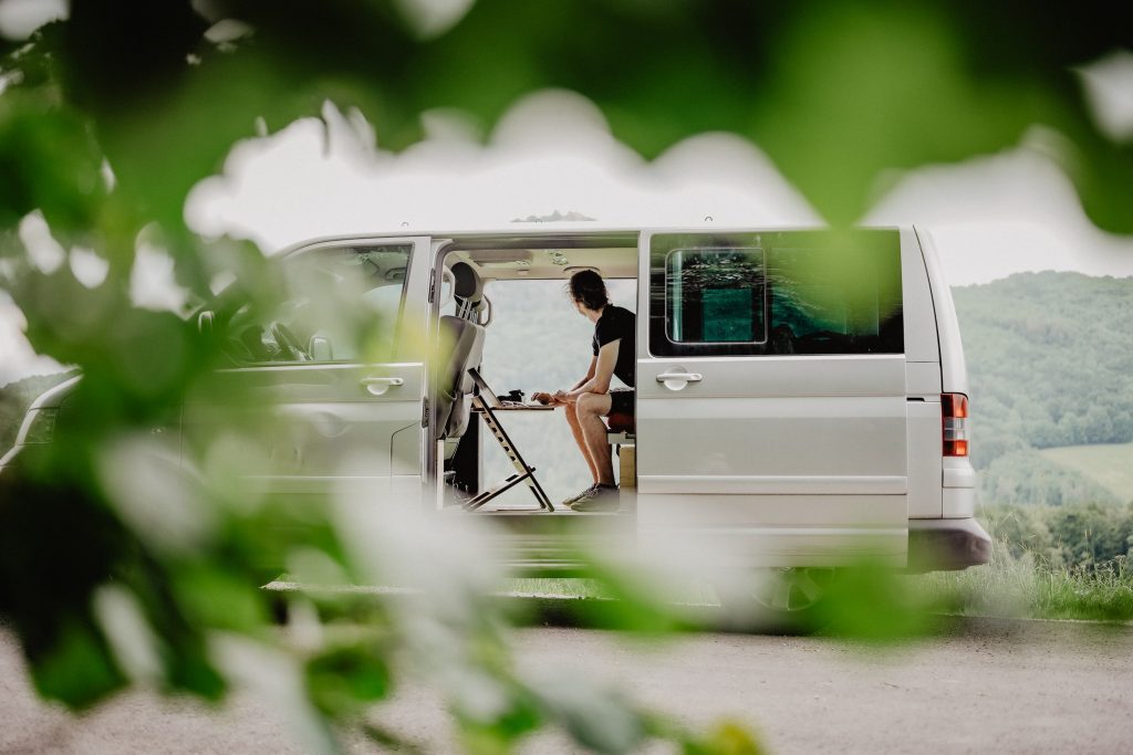 A man sitting in a van. In the foreground we have an out of focus bush framing him neatly. This man is clearly at a stopping point along his van life journey.