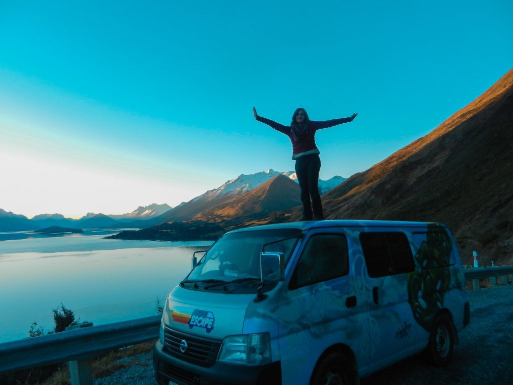 Cornish and cosy co-founder Becca standing on a van during travels in New Zealand. There is a deep blue sky that falls into snow capped mountains. Becca and her friends have been living the van life for a few months as they explore the beautiful country of New Zealand.