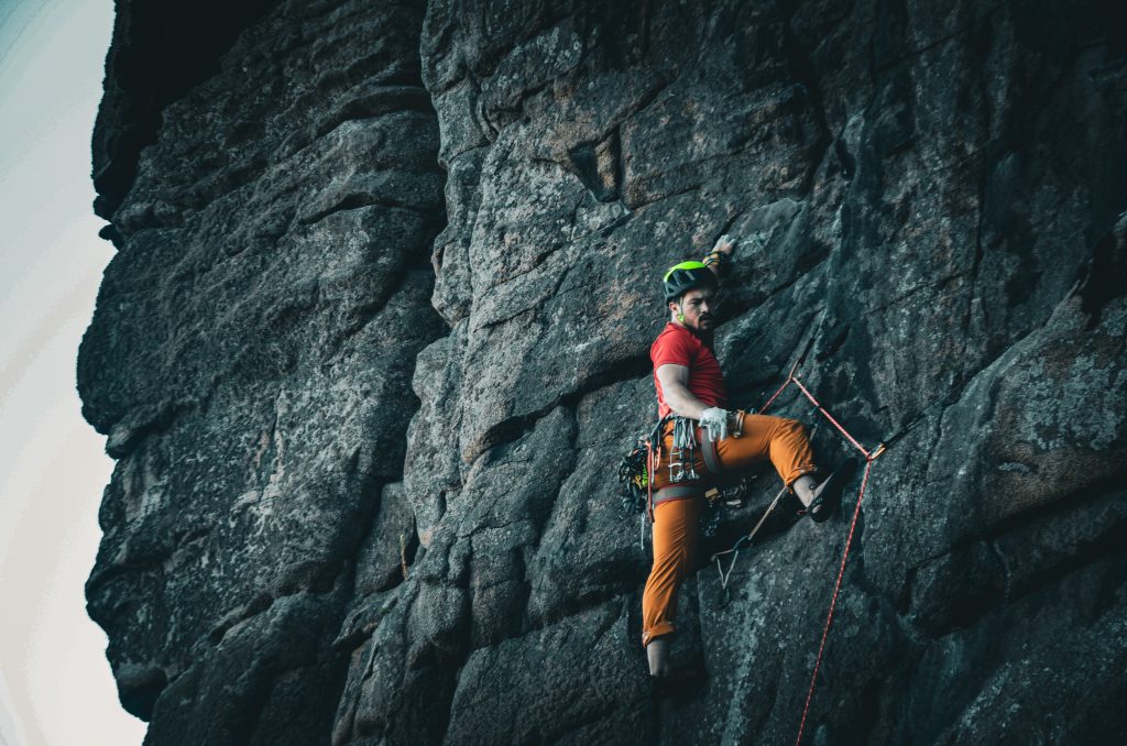 A man can be seen climbing. He is wearing bright red and orange and climbs in a harness and helmet. He is clinging on to the cliffside in Cornwall during one of his adventures. 