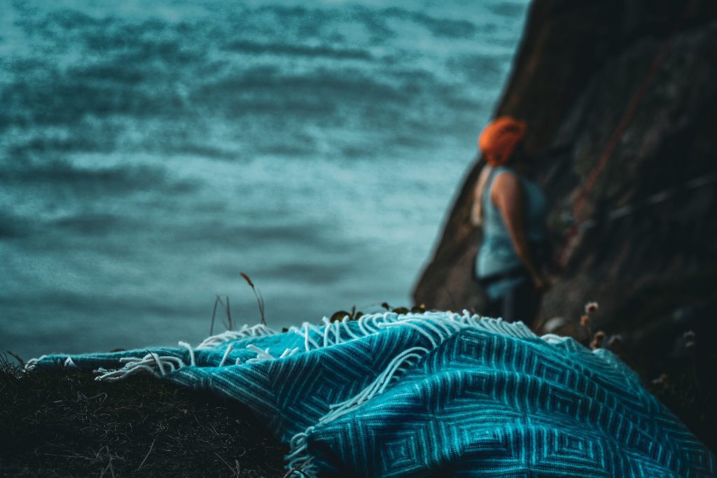 A blanket sits upon a cliff looking out to sea. A woman is in the background belaying as a climber climbs up a cliffside. 
