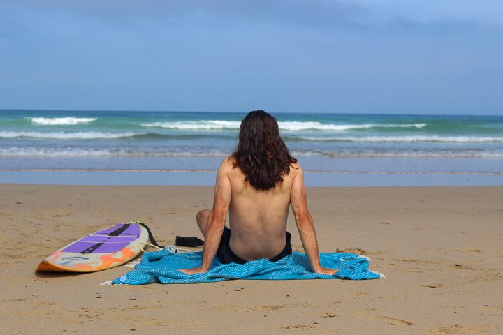 A man sits atop a bright blue blanket whilst looking out to sea. He sits next to his surfboard and appears to have just been for a surf. The waves are calm and the sea is blue. The beach is in Cornwall, the man's home. 