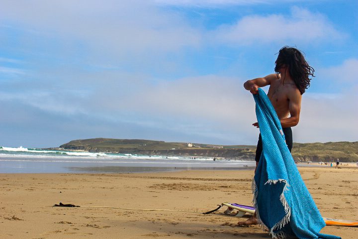 A young man, near his home in Cornwall, stands on a beach looking out to sea. he has just been for a surf and is now about to wrap up warm in a blanket. It is a sunny day. You can see waves in the background. 