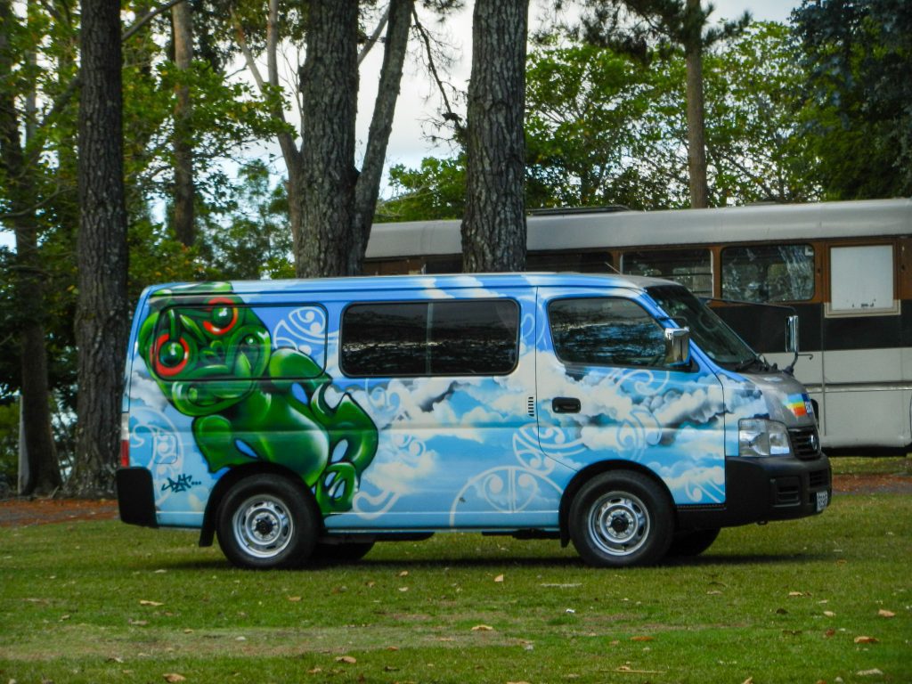 A photograph of a campervan. The Campervan is painted in a blue and white pattern with a tike cartoon which is green with red eyes. The van is parked near another van in a campsite with lots of tall trees and grass. The van is travelling through New Zealand. 