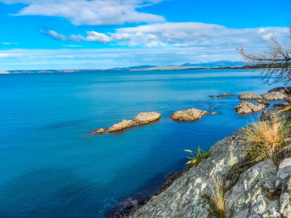 A photograph taken in New Zealand of a beautiful blue ocean. The foreground is rocky and grassy. There are rocks in the ocean. There are lots of mountains in the background. It is a sunny day. The view can be scene when travelling through New Zealand by van. 