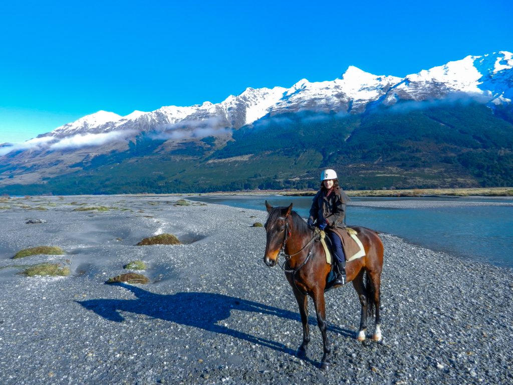 A photograph of a young woman riding a big bay horse. They are near a long river with bright blue water. The river is below a snow-capped mountain range. It is a sunny day. The photo was taken in Queenstown, New Zealand. Horse riding is a great way to travel. You can find lots of spots to horse ride by van. 