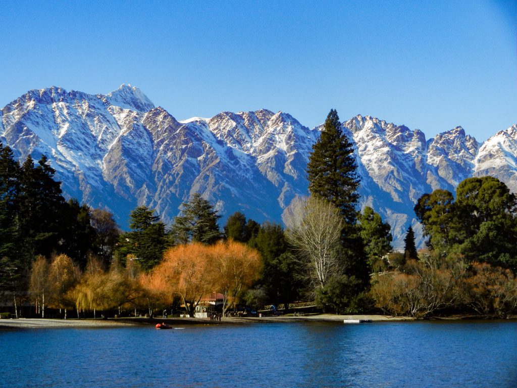 A photograph of a snow-capped mountain landscape. The mountains are beautiful and grey and blue in colour. There are lots of green trees lining the base of the mountain and a blue lake. The photograph was taken in Queenstown, New Zealand. Queenstown is very van-friendly and good for travel. 