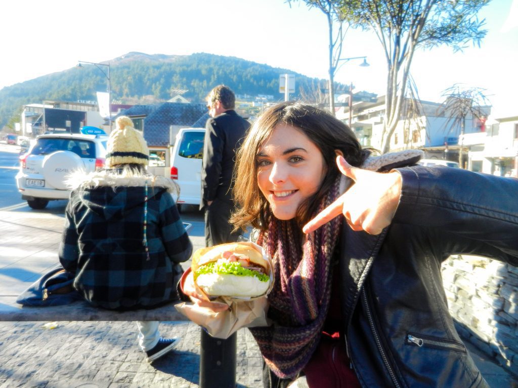 A photography of a young woman proudly holding a very large beef burger and pointing to it. There are people in the background and a mountain. The person has stopped to refill after a long-day of driving the van. 