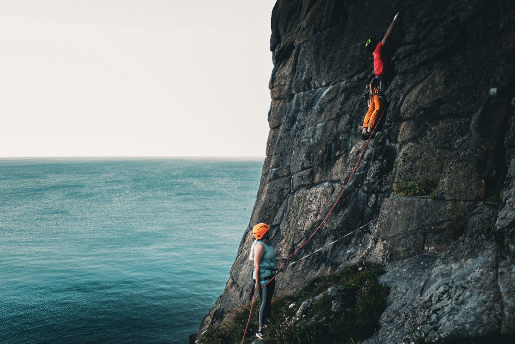 A man and woman can be seen climbing. The man is climbing the cliff while the woman belays from below. The ocean is in the background. They are in Cornwall on an adventure. 