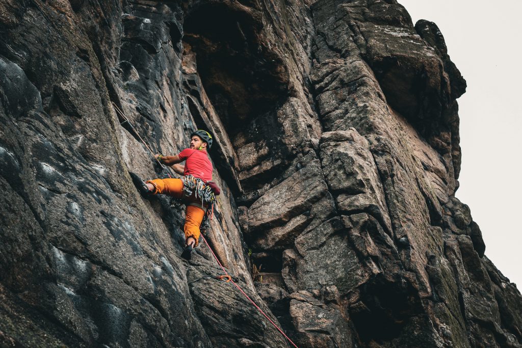 A man can be seen climbing. He is wearing bright red and orange and climbs in a harness and helmet. He is clinging on to the cliffside in Cornwall during one of his adventures. 