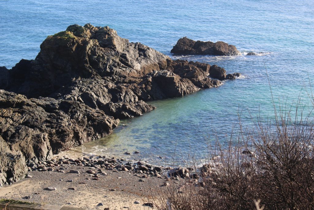 A photo of the ocean and a small, rocky beach in Cornwall. It is a sunny day. This a is great place for a hike. 