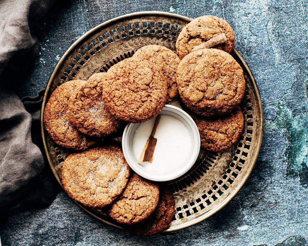 A picture of a plate of ginger biscuits. They are a brown, spiced colour and in the centre is a pot of spice milk with cinnamon. They are made from a traditional Cornish recipe and are perfect for picnics. 