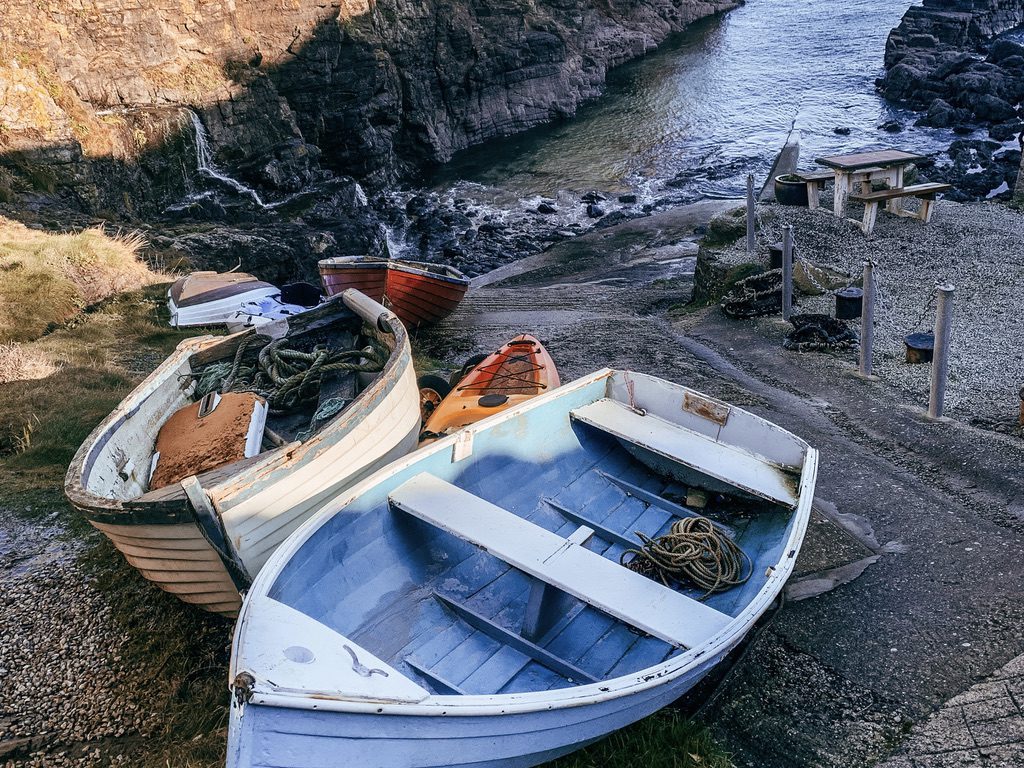 A photo of some small and colourful boats in a small fishing harbour in Cornwall. This is a stopping point on a hike. The waters of the sea are blue in the background. 