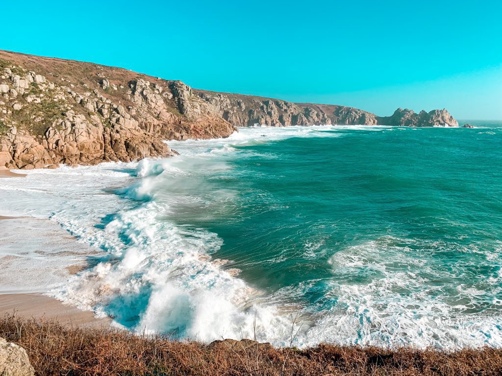 A photo of Porthcurno, a a beach in Cornwall. The photo is taken from the Minack theatre and you can see the coastline. The sea is turquoise and the waves crash against the beach. 