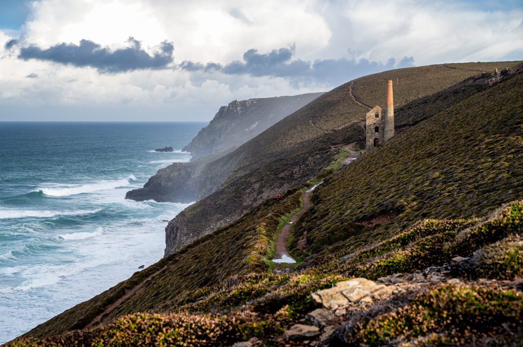A photograph of Wheal Coates, a mining engine in Cornwall. The photo shows the coast path which is great for hiking down t the beach. It's a sunny day and the waves are crashing against the cliffs. 