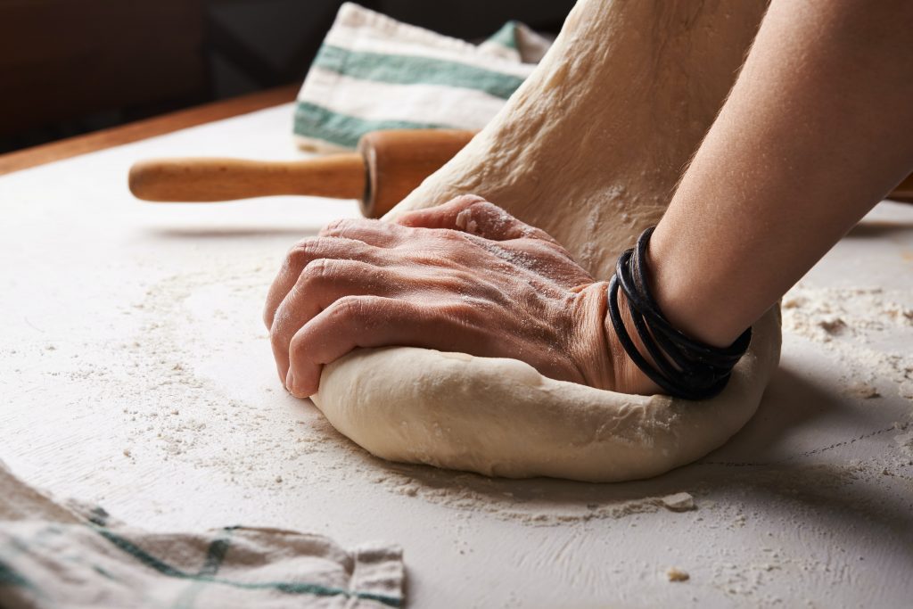 A photography of a woman baking bread. She is kneading the dough with one hand and is covered in flour. She is baking a traditional Cornish recipe for her family when they go on a picnic. 