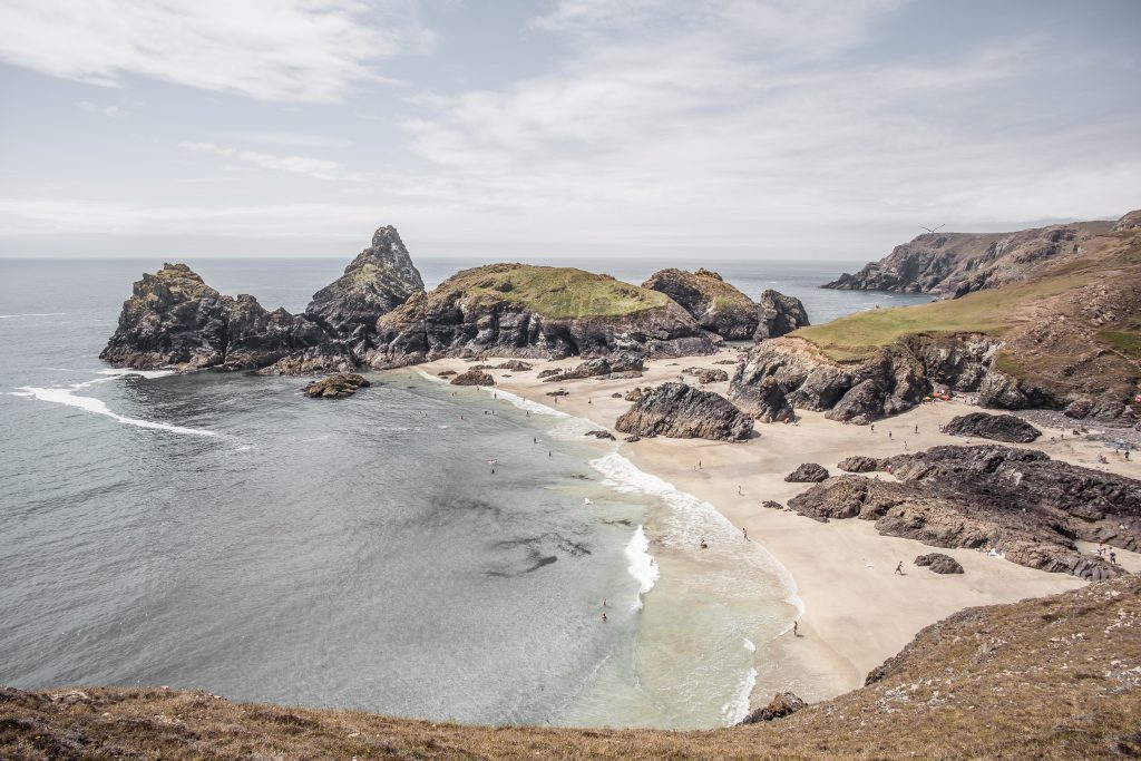 A photo of Kynance Cove in Cornwall. The start of the hiking trail on the coast path. It's a wintry day, the tide is out and the beach is large with some people walking on it. 