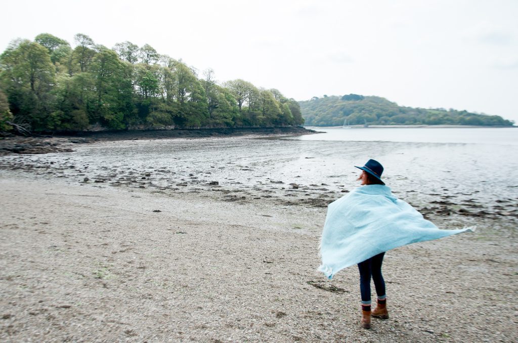 A photo of a young woman wrapped in a blue blanket whilst walking along a beach. The beach is on a river and there are trees in the background.