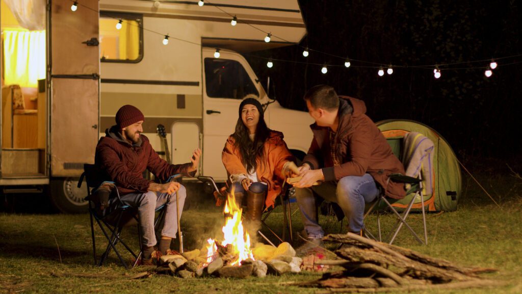 Group of close friends laughing together around camp fire. Retro camper van. Light bulbs in the background.