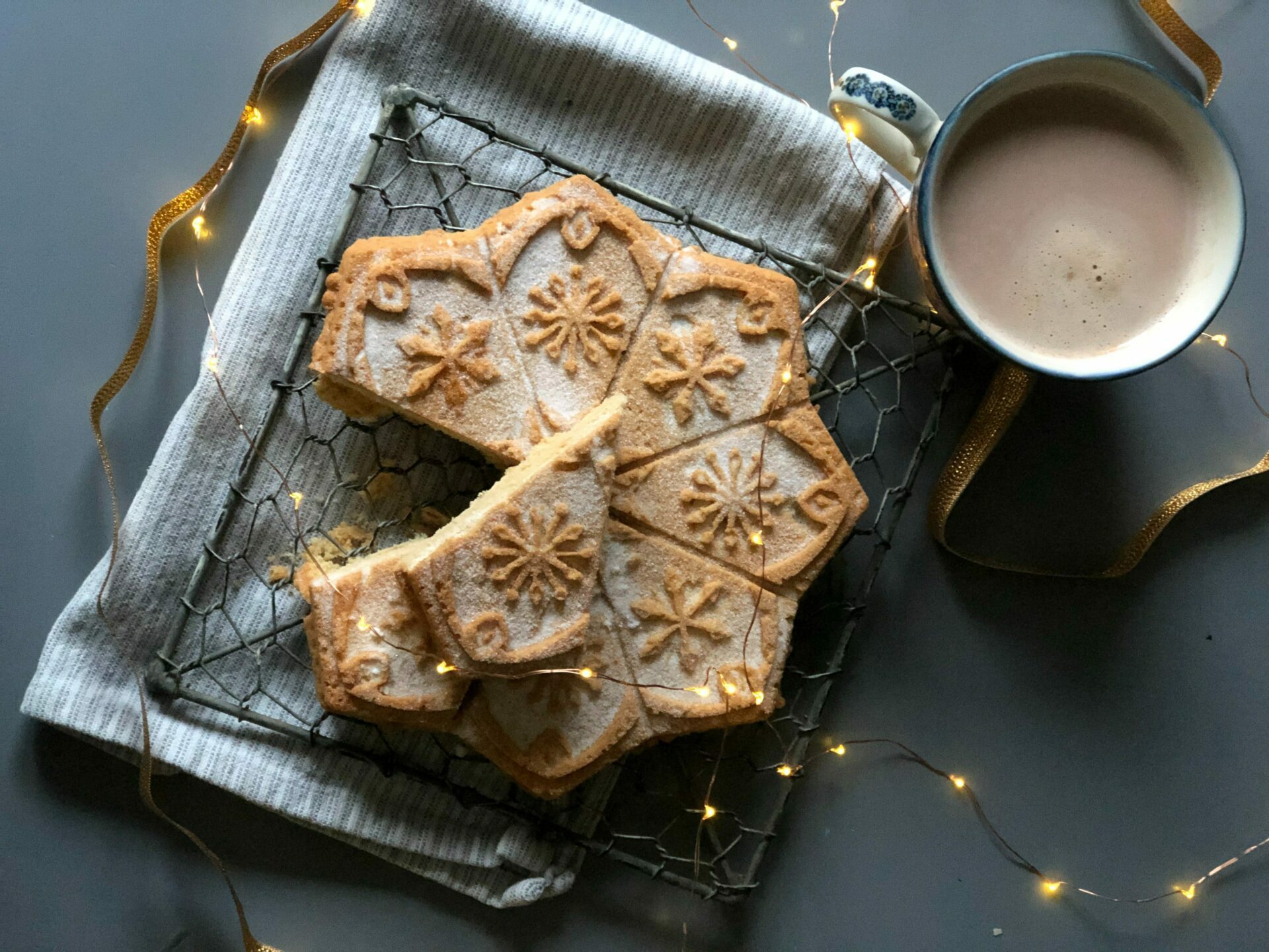 A photograph of a mug of hot chocolate and sugary shortbread. Fairylights surround them. 