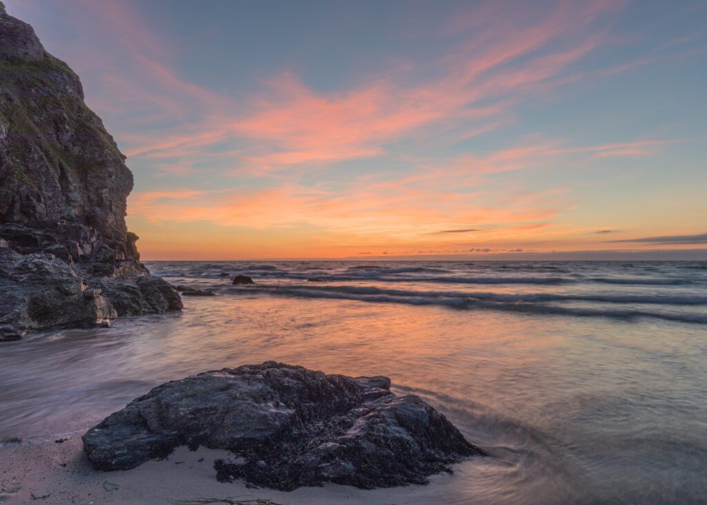 Cosy sunset at Porthtowan. Cliffs on the left, rock in the foreground with oranges and pinks in the sky over the sea.