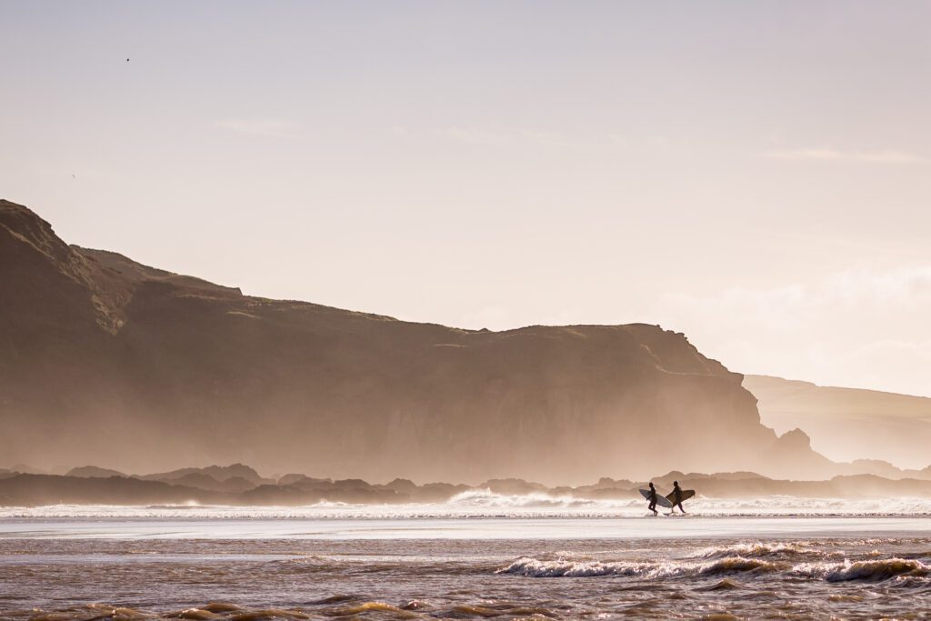Image of two surfers in the sea. Walking back to shore with dramatic cliffs behind them.