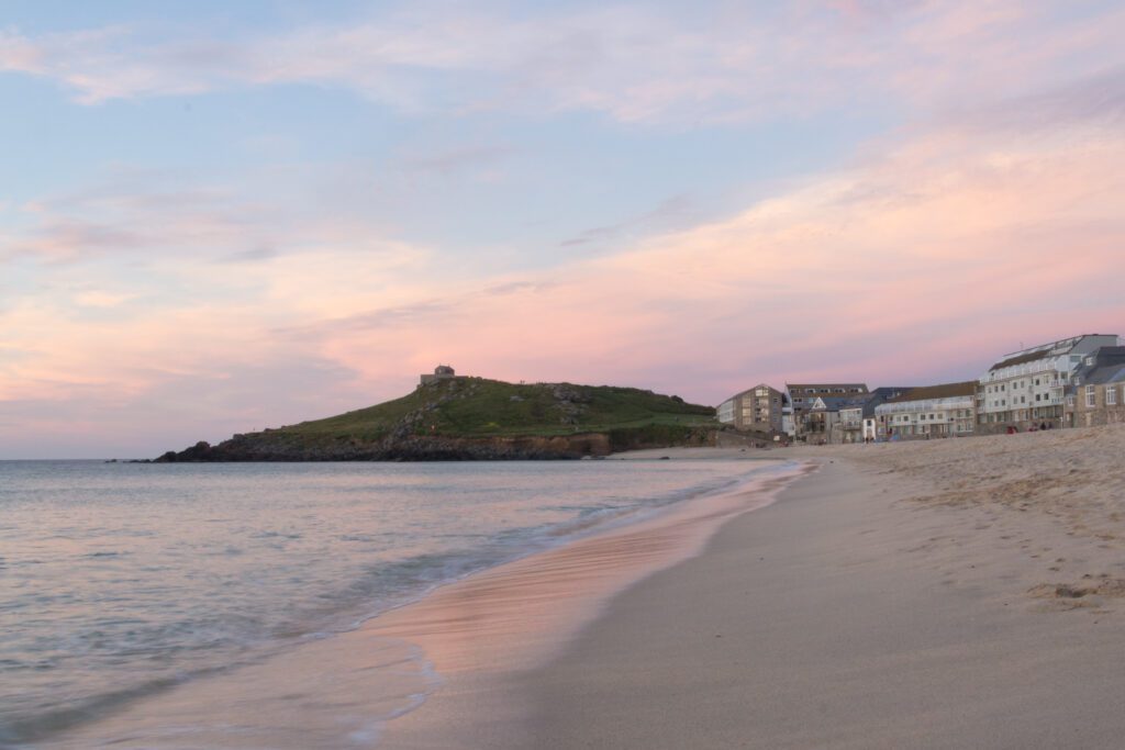 Cosy sunset at Porthmeor. Shoreline in the foreground with buildings on the right in the background and the headland on the left. Sky is filled with pink and purple colours.