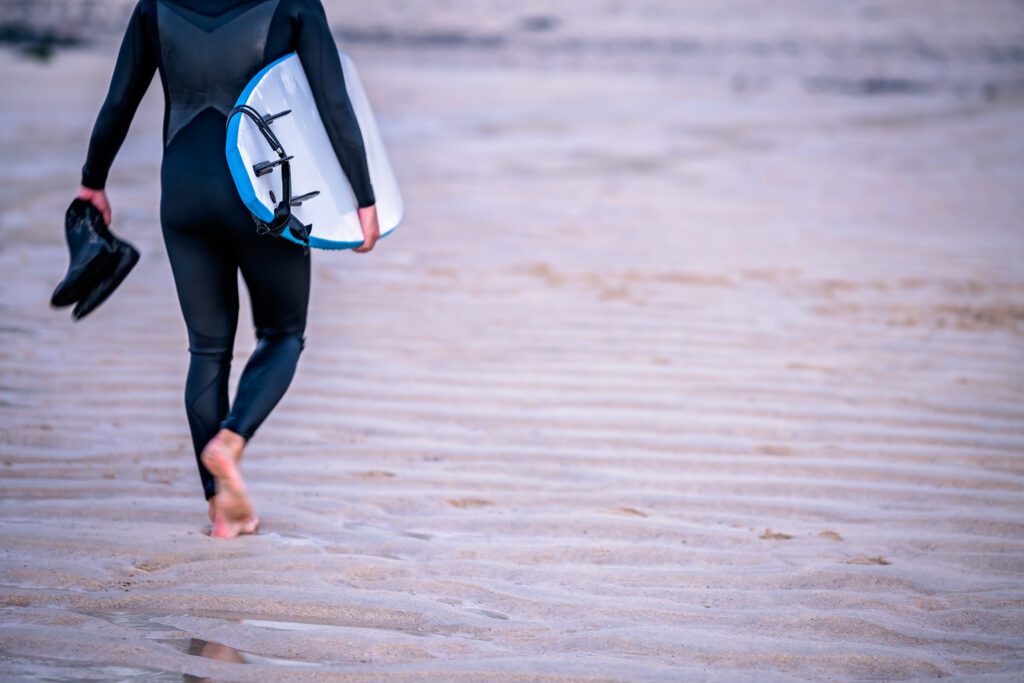 A man walking to the surf. He has his boots in one hand, surf board in the other walking along the sand