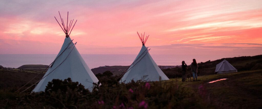 Sunset over the tipi tents at Kudhva. There are 3 tents visible with the sea in the background. The sky is electric pink as the sun dips behind the water.